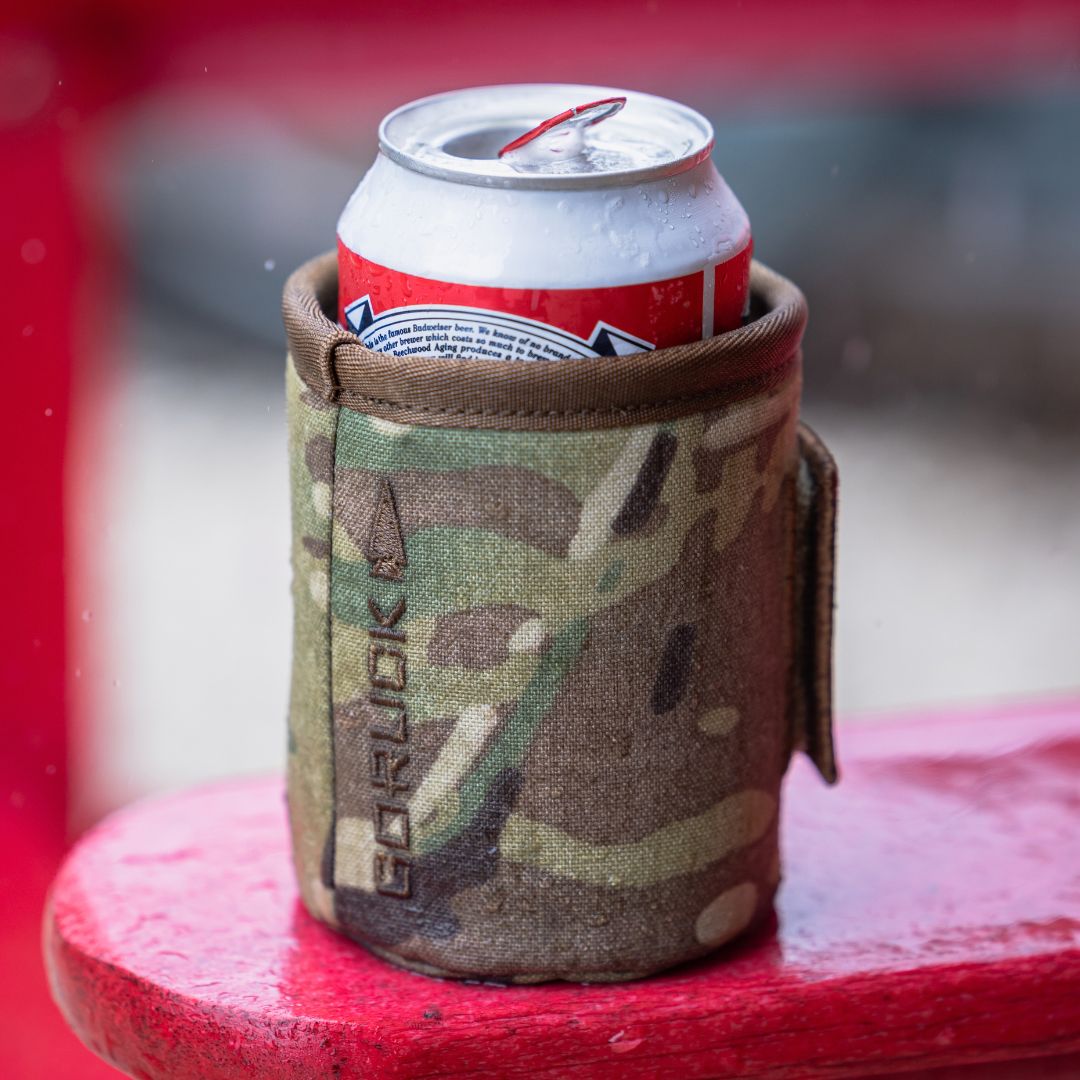 A camo-print Ballistic Beer Jacket holding a can of beer on a red surface.