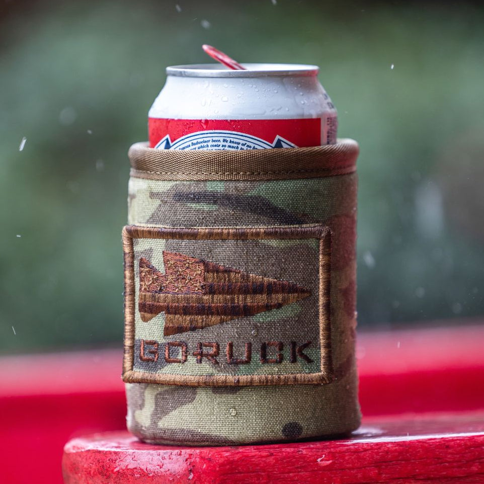 A camouflage Ballistic Beer Jacket with a GORUCK spearhead patch sits on a red surface in the rain.