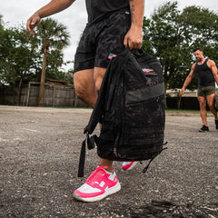 A person wearing pink and white sneakers carries a large black GORUCK Rucker 4.0 backpack, strolling outdoors on a gravel path. Another individual in dark athletic clothing follows behind, rucking through the area with trees and a fence visible in the background.