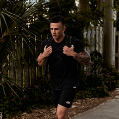 A tattooed man in black workout clothes carries a Ruck Plate Carrier 3.0, made from ballistic nylon, while walking outdoors.