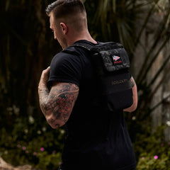 Man with a flower tattoo on his arm wears a black Ruck Plate Carrier 3.0 backpack made from ballistic nylon, enjoying the outdoors.