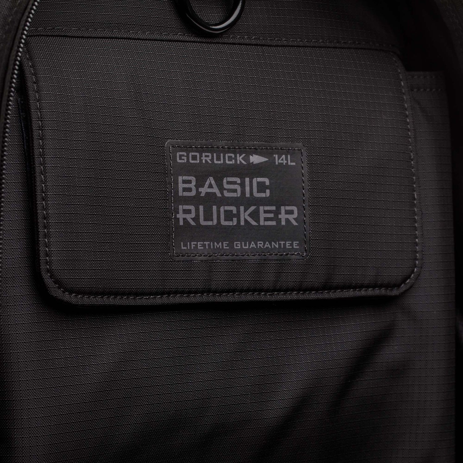 Close-up of the Basic Rucker backpack in black, showcasing its pocket and label reading 