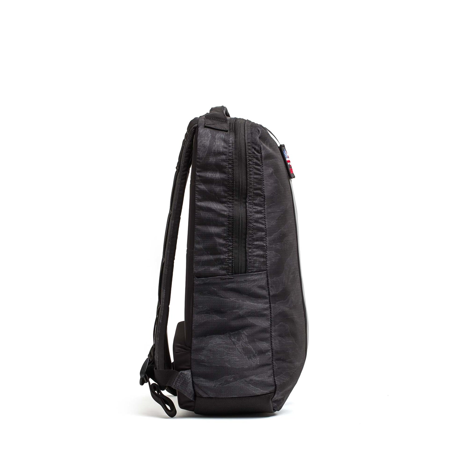 Side view of the Basic Rucker, a black backpack with a zipper and adjustable shoulder straps, shown against a white background.