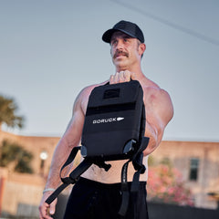 Outdoors, a shirtless man wearing a black cap holds the Ruck Plate Carrier 3.0 backpack at arm’s length, highlighting its built-in lumbar support.
