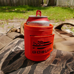 A red beverage can fits perfectly in an orange Ballistic Beer Jacket - GORUCK, resting on a camouflage backpack crafted from durable 1000D CORDURA, with grass and a fence in the background.