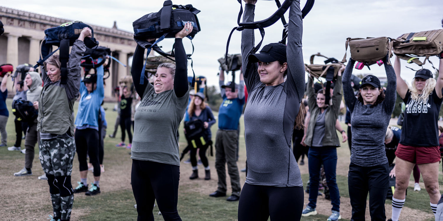 A group of people outdoors lifting sandbags overhead during a fitness workout on grass.