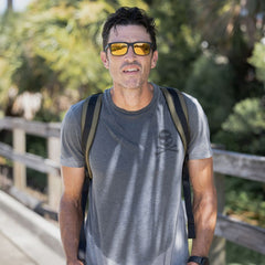 Wearing the GORUCK Tribe Tee - Tri-Blend and yellow sunglasses, a man with a backpack stands outdoors on a sunny day, ready to tackle the next GORUCK Tribe challenge as part of a supportive community.