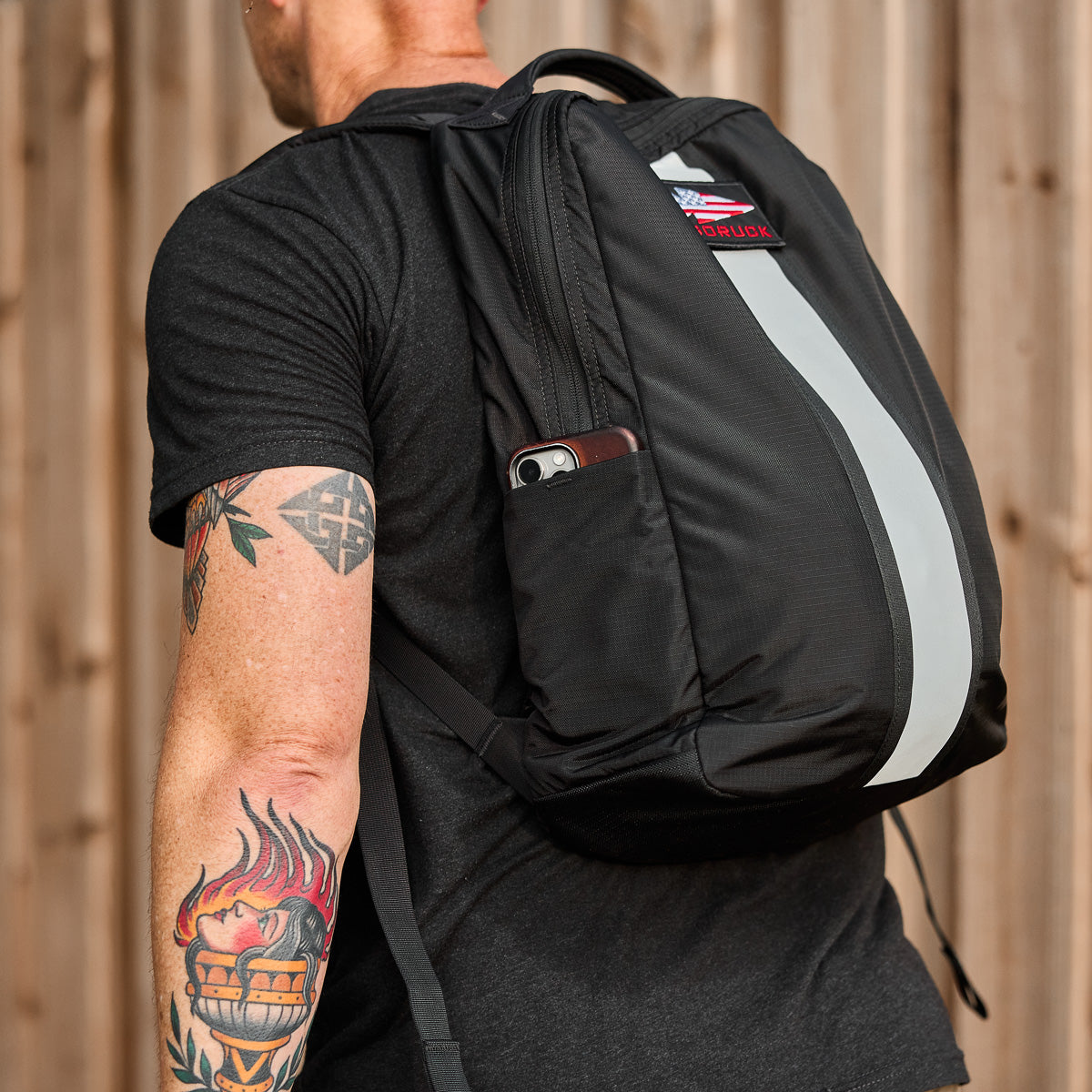 Man with tattoos wearing a black backpack with a reflective stripe and a phone in the side pocket.