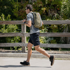 A man jogs on a sunny day, wearing sunglasses, a backpack, athletic clothes, and comfortable Merino Challenge Socks - Ankle (No-Show), with greenery in the background.