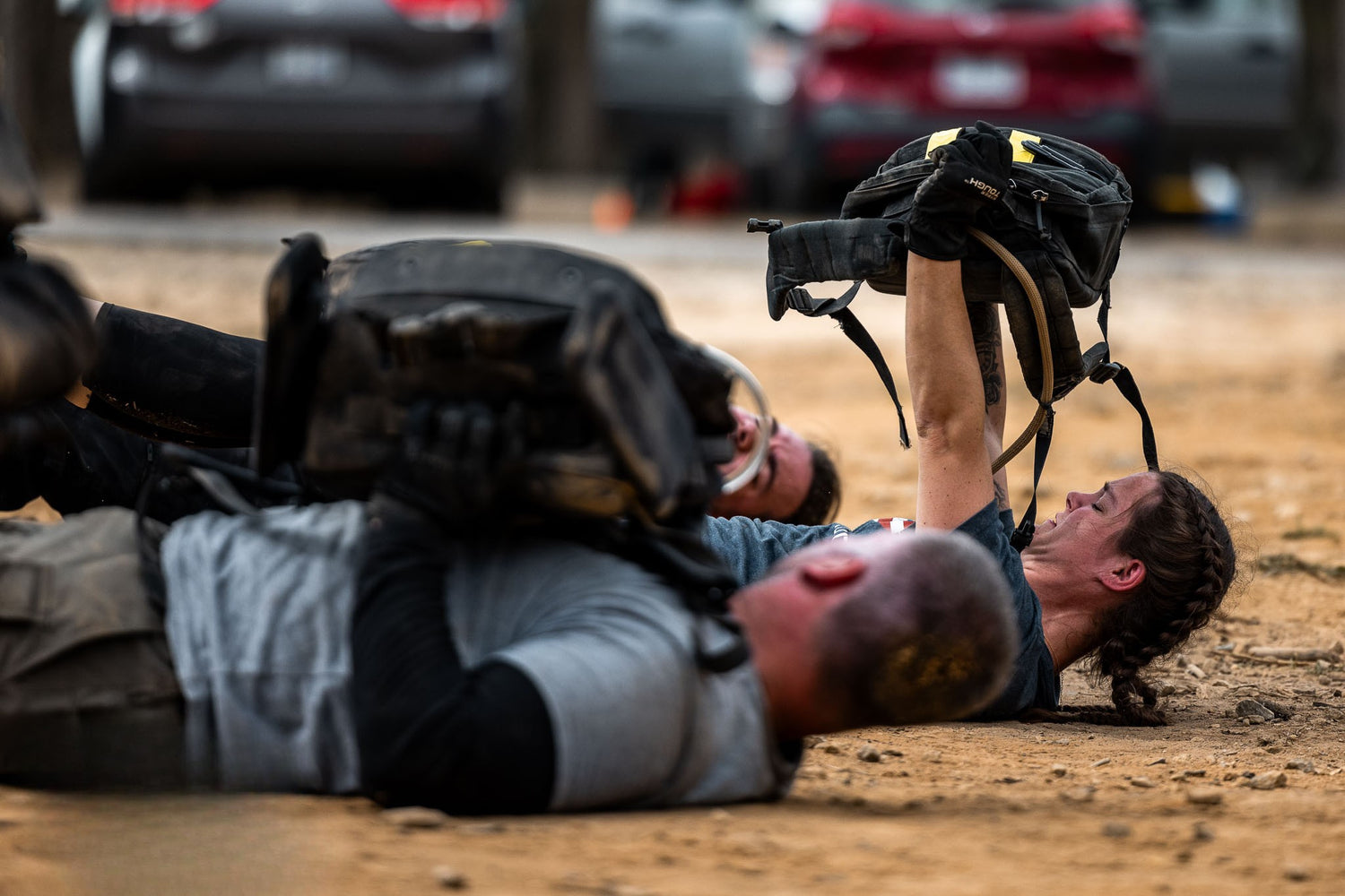 Two people lying on the ground outdoors, lifting backpacks above their chests during a workout.