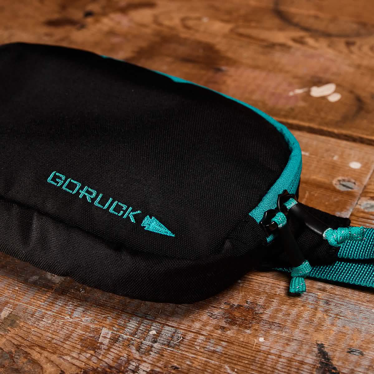 On a textured wooden surface sits a compact Belt Bag from GORUCK in a sleek black color with teal accents. Constructed from durable Cordura fabric, the bag showcases a prominent zipper and a strap adorned with teal details. The GORUCK logo, embroidered in teal, stands out on the bag.