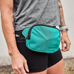 A person wearing a gray t-shirt and black shorts is sporting a teal GORUCK Belt Bag with adjustable webbing to keep their gear organized. The bag features a zipper and a small logo, while tattoos embellish the person's right arm, complemented by a smartwatch on their wrist.