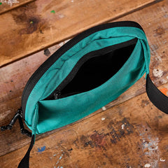 The GORUCK Belt Bag in teal fabric, featuring a black zipper, is open to show its compact interior. Positioned on a paint-splotched wooden surface, the bag includes adjustable webbing and a black strap attached to one side.