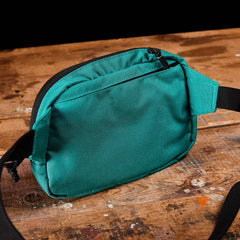 A small, green fabric GORUCK Belt Bag with black adjustable webbing and zipper rests on a rustic wooden surface. This organized piece boasts a straightforward design featuring a front pocket for added convenience.