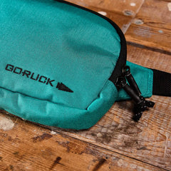 A GORUCK Belt Bag, featuring a teal color with black zippers and straps, is placed on a wooden surface. The bag includes adjustable webbing for perfectly organized gear, with the brand logo prominently displayed.
