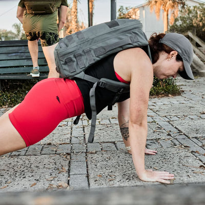 A person is doing push-ups outdoors, wearing a cap and red Women's Biker Shorts made from ToughFlex fabric by GORUCK. Their backpack intensifies the challenge, reflecting resilience and endurance in the open air.