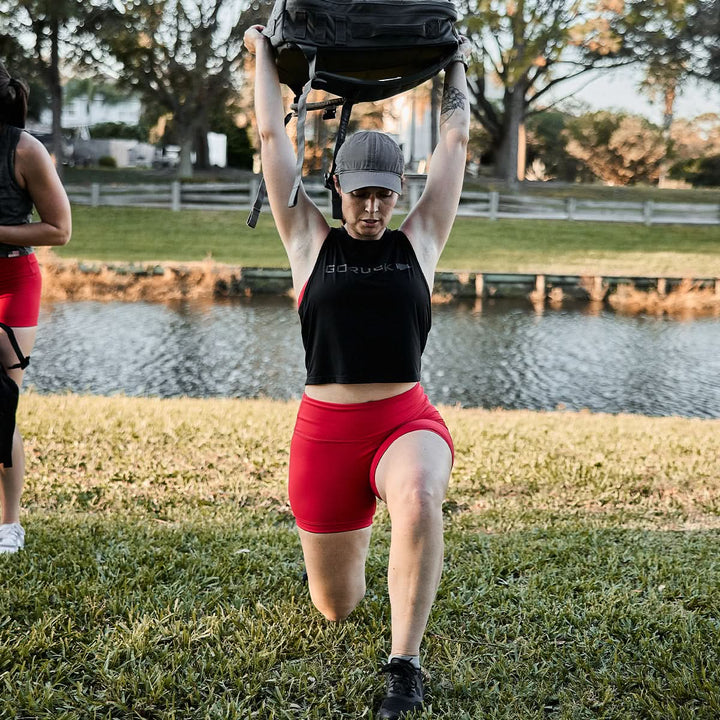 A woman in ToughFlex biker shorts by GORUCK lunges outside, lifting a weighted bag overhead near a pond.