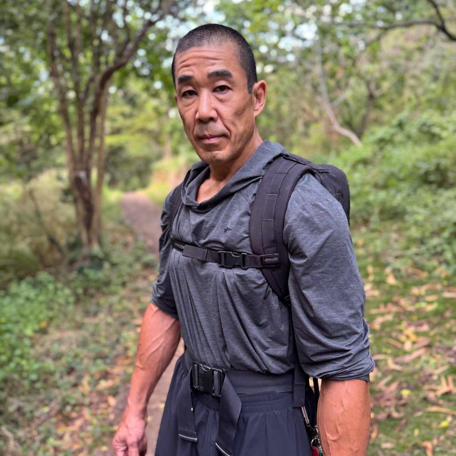 A man wearing a gray shirt and backpack stands on a wooded trail surrounded by greenery.
