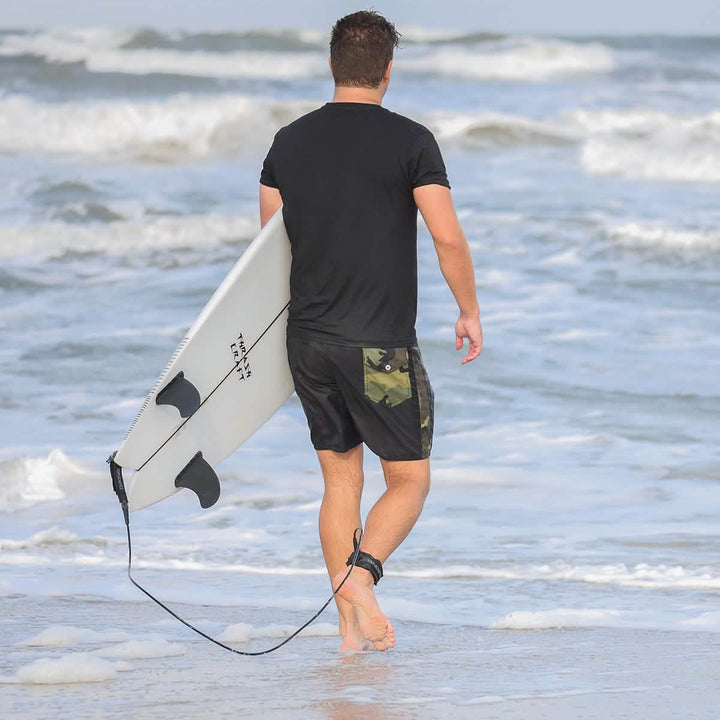 A man strolls along the beach, holding a surfboard under his arm. He wears GORUCK's 300 Boardshorts with a camouflage pattern, paired with a black shirt. The waves crash in the background, and a surfboard leash is secured to his ankle.