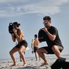 Two individuals squatting on a beach holding black GORUCK sandbags during outdoor workout