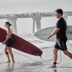 Man and woman carrying surfboards on beach near pier, both wearing black board shorts