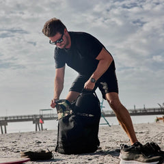 Man in black outfit packing a GORUCK backpack on a sandy beach with a boardwalk in the background