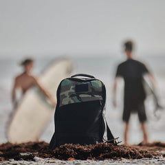 Black and camo GORUCK backpack on sandy beach with two surfers holding surfboards in background