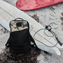 GORUCK tactical backpack with camo top resting on sandy beach next to surfboards and leash