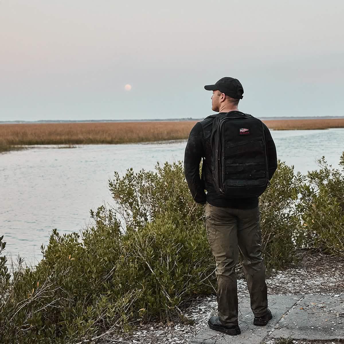 A person stands on a path near water, wearing a ToughDry® fabric black jacket and a GORUCK x Birdwell Tac Hat w/Patch, with a backpack, gazing at the horizon. The scene includes bushes and a scenic view of grassy marshland under an overcast sky.