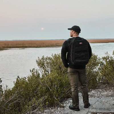 A person stands on a path near water, wearing a ToughDry® fabric black jacket and a GORUCK x Birdwell Tac Hat w/Patch, with a backpack, gazing at the horizon. The scene includes bushes and a scenic view of grassy marshland under an overcast sky.