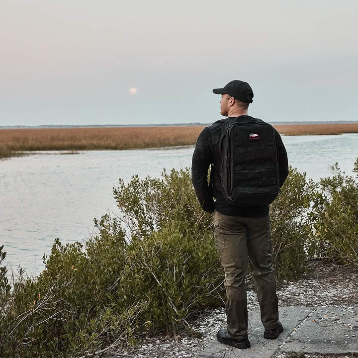 A person stands on a path near water, wearing a ToughDry® fabric black jacket and a GORUCK x Birdwell Tac Hat w/Patch, with a backpack, gazing at the horizon. The scene includes bushes and a scenic view of grassy marshland under an overcast sky.