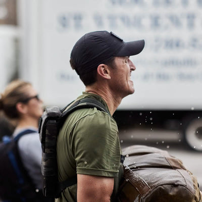 A man wearing the GORUCK x Birdwell Tac Hat w/Patch, which is black and complemented by a green shirt, hauls a heavy sandbag through splashing water, exemplifying the durability required of GORUCK gear. In the background, another individual fitted with sunglasses and a backpack emphasizes his exertion-driven task.