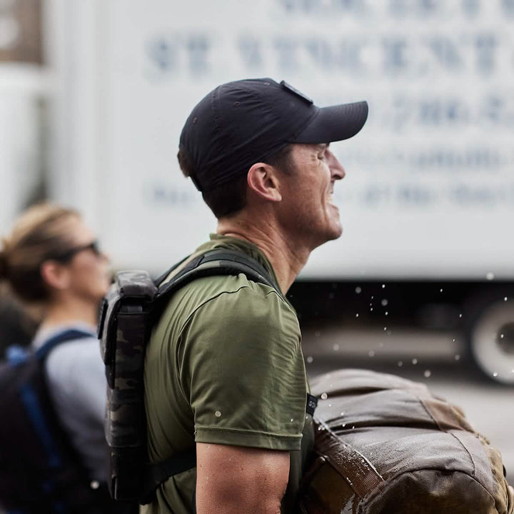 A man wearing the GORUCK x Birdwell Tac Hat w/Patch, which is black and complemented by a green shirt, hauls a heavy sandbag through splashing water, exemplifying the durability required of GORUCK gear. In the background, another individual fitted with sunglasses and a backpack emphasizes his exertion-driven task.