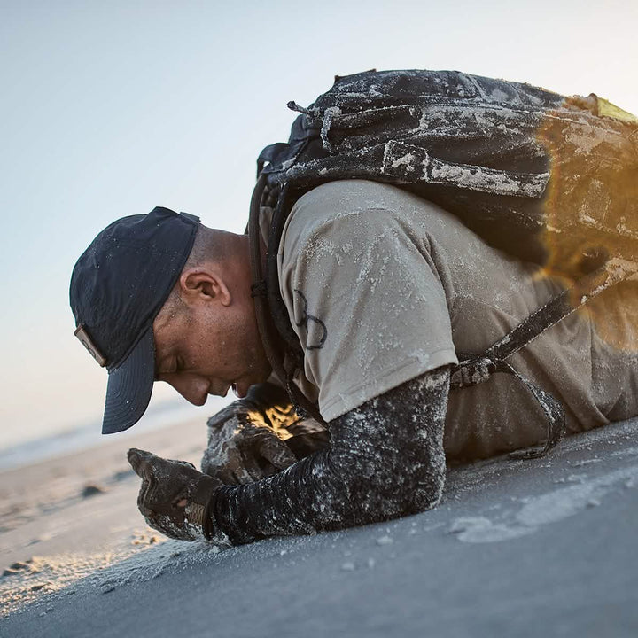 A person wearing a GORUCK x Birdwell Tac Hat w/Patch and backpack lies face down on a sandy beach, their clothing dusted with sand. The sun sets in the background, casting a warm glow. They appear focused, possibly examining something on the ground made from ToughDry® fabric.