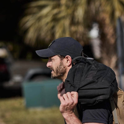 A man in a black GORUCK x Birdwell Tac Hat w/Patch and carrying a black backpack strolls outdoors, surrounded by swaying palm trees. His gear is crafted from ToughDry® fabric, epitomizing the durability and style of the GORUCK x Birdwell collaboration.