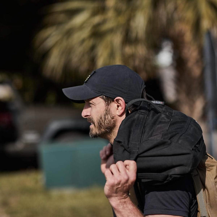 A man in a black GORUCK x Birdwell Tac Hat w/Patch and carrying a black backpack strolls outdoors, surrounded by swaying palm trees. His gear is crafted from ToughDry® fabric, epitomizing the durability and style of the GORUCK x Birdwell collaboration.
