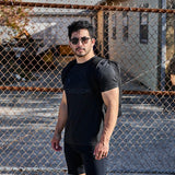 Man wearing black GORUCK t-shirt and backpack outdoors by a chain-link fence