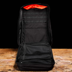 A black GR2 tactical backpack from GORUCK is displayed open on a wooden surface against a dark background. It reveals multiple interior compartments, mesh pockets, and vibrant orange lining - perfect for Special Forces training or everyday ruck adventures.