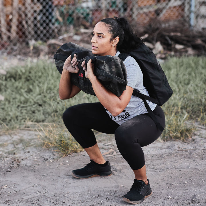 A woman squats outdoors with a sandbag on her shoulders, wearing a gray shirt and black Women's Cropped Training Leggings. Made from ToughFlex fabric, these durable leggings offer comfort and are squat-proof for intense workouts.