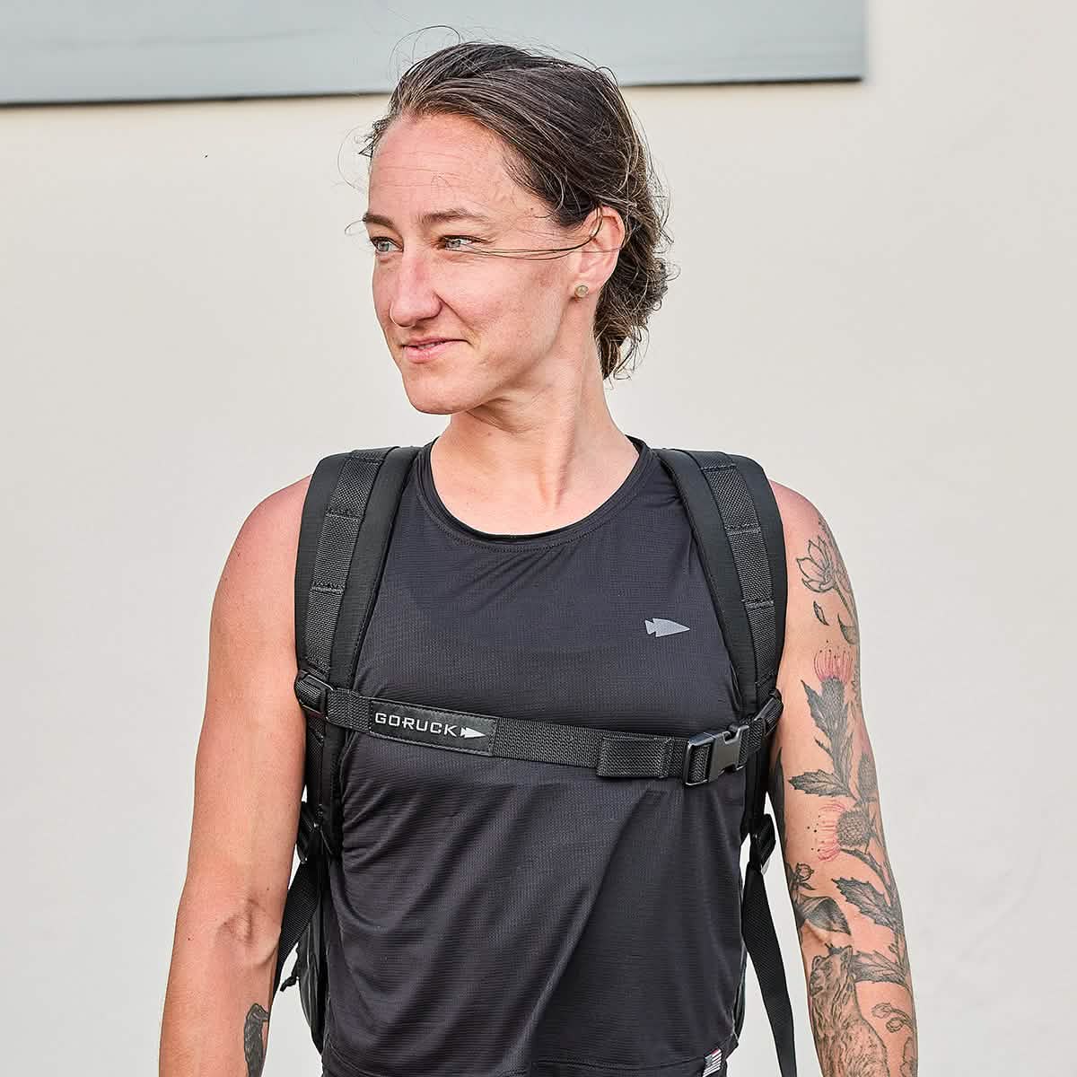 A woman in a black tank top with a compact GR0 - Ripstop ROBIC® rucksack stands outdoors, looking to the side and displaying a tattoo on her arm.