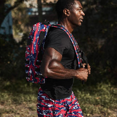 Athletic man wearing black shirt and red-blue camo shorts carrying colorful rucking backpack outdoors