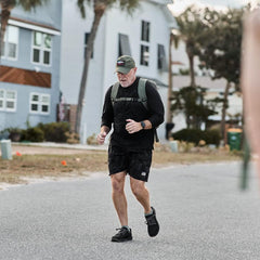 Middle-aged man running outdoors in black athletic gear, wearing a backpack and cap in a residential neighborhood