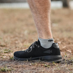 Side view of a person wearing black GORUCK rucking shoes and gray ankle socks outdoors on dry grass