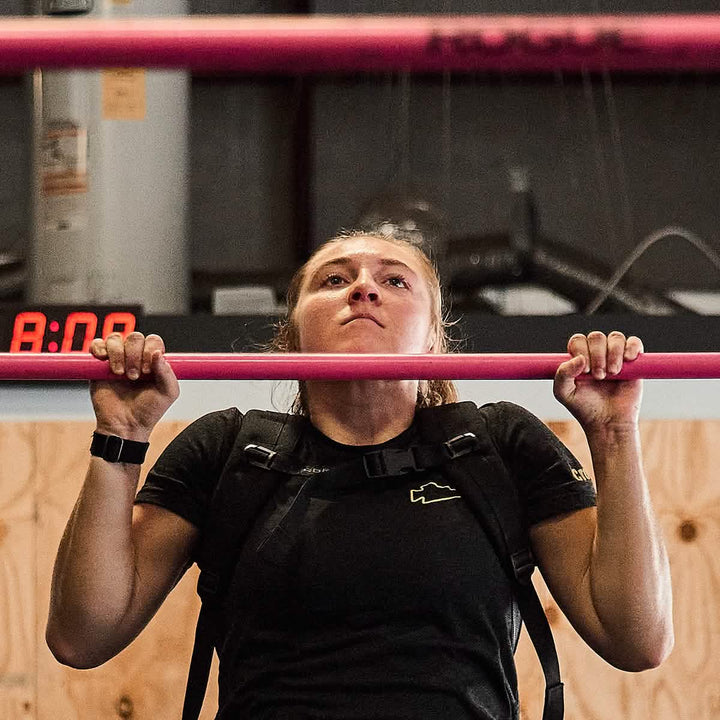 Wearing a GORUCK Women's Subtle Spearhead x CrossFit Cropped Tee in premium poly-blend, paired with a weight vest, an individual executes pull-ups on a pink bar. The backdrop comprises a wooden wall and industrial features, with the red digital clock displaying 8:00 to emphasize the intensity of this workout session.