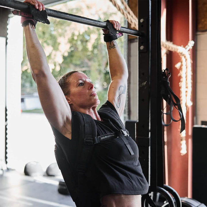 In a sunlit gym, a person dressed in athletic gear and wearing the GORUCK Women's Subtle Spearhead x CrossFit Cropped Tee - Poly-Blend executes a pull-up with a weighted vest. Their intense focus underscores the dedication of their workout surrounded by gleaming equipment.