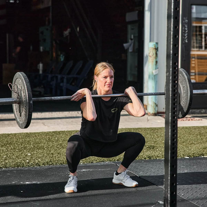 A person confidently performs a front squat with a barbell in an outdoor gym, wearing the stylish Women's Subtle Spearhead x CrossFit Cropped Tee - Poly-Blend by GORUCK and leggings paired with white sneakers. The background reveals workout equipment and a grassy area bathed in bright sunlight, highlighting their dedication.