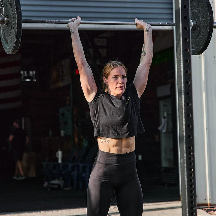 In a GORUCK Women's Subtle Spearhead x CrossFit Cropped Tee – Poly-Blend, a person lifts a barbell above their head in an outdoor gym. Sunlight highlights their toned muscles and tattoos, while the premium poly-blend fabric of the tee enhances comfort during the workout.