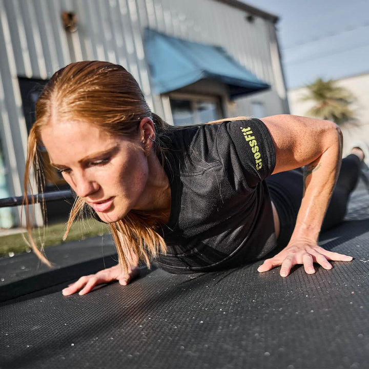 A red-haired woman is performing a push-up on a black exercise mat outdoors, wearing the Women's Subtle Spearhead x CrossFit Cropped Tee by GORUCK. Her dedication to her workout stands out against the backdrop of a building and tree.