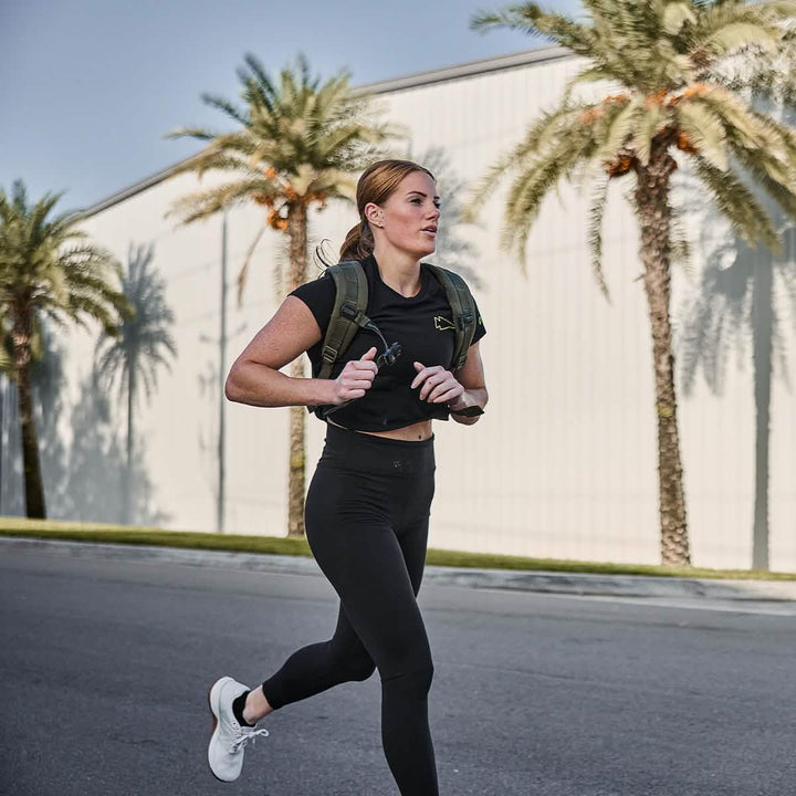 A person jogging on a palm tree-lined street, sporting a black Women's Subtle Spearhead x CrossFit Cropped Tee by GORUCK, along with white sneakers and a green backpack. A large building stands in the background under a clear blue sky.