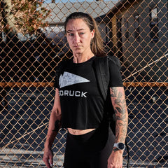 In front of a chain-link fence, an individual stands confidently, sporting a Women's Cropped Tee - Poly-Blend by GORUCK in black. They pair it with a smartwatch and backpack, showcasing tattoos on one arm against an urban backdrop of buildings and trees while looking at the camera.